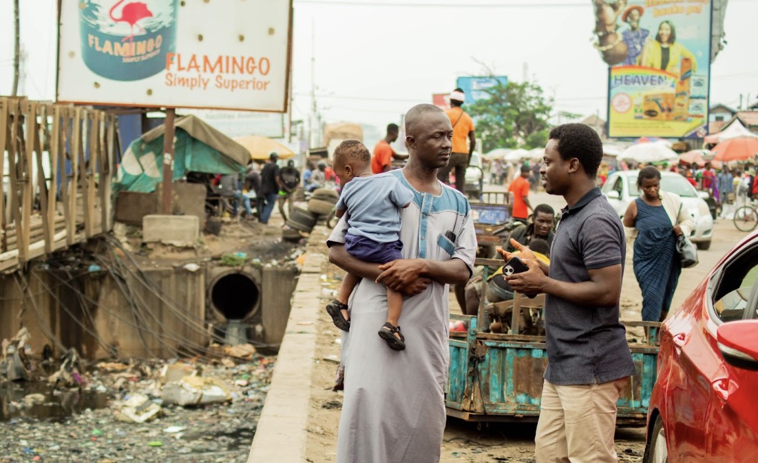 Albert Oppong Ansah interviewing a man carrying a child in a Ghanaian market.